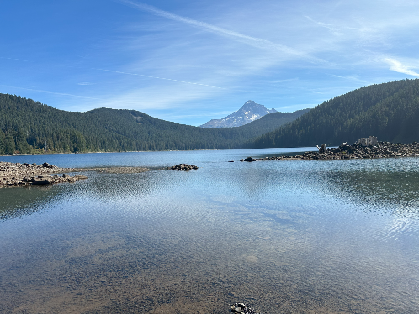 blue lake surrounded by rolling, forested foothills with mount Hood in the background. Blue sky day.