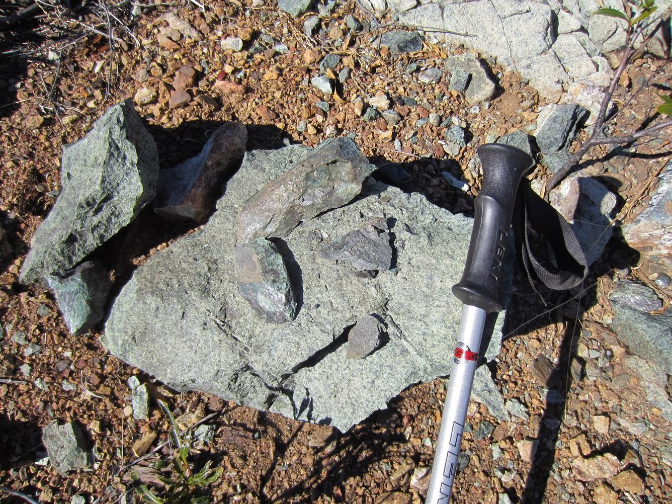 A greenish boulder with several shiny green rock fragments rests on pebble-strewed ground. The boulder is mottled with several colors of green minerals. The grip end of a hiking pole rests on the boulder to give a sense of scale. 