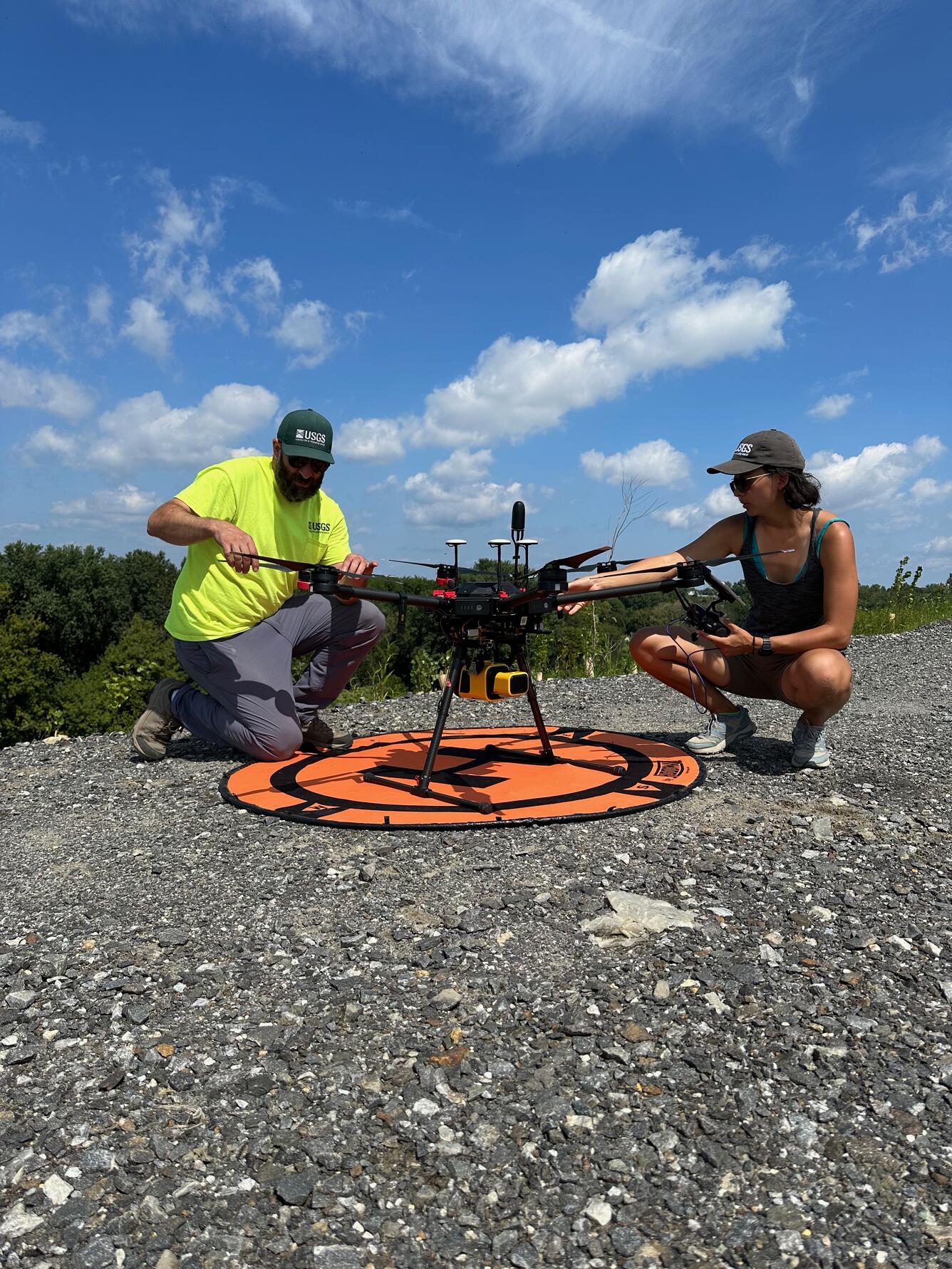 Two people kneeling on ground next to a drone on a sunny day