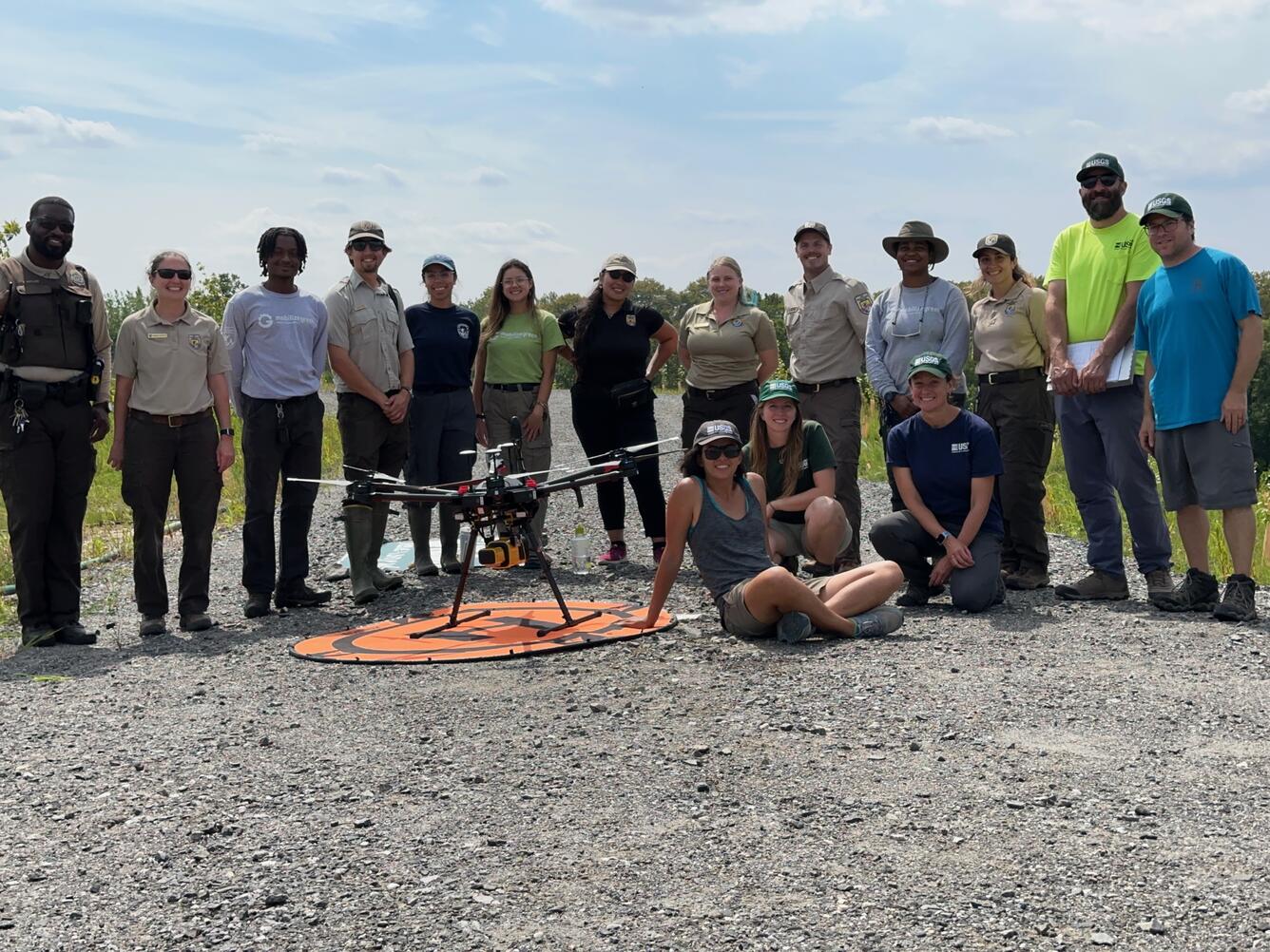 Large group of people outside smiling for photo with drone on the ground in front of them