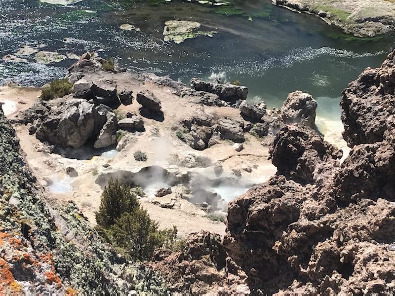 Looking over the edge of a rocky cliff, a packed dirt creekside is scattered with boulders of volcanic rock. In the middle of the bare dirt, a string of four circular sinkholes bubble with frothing water. Steam rises above the holes and obscures their surfaces. USGS photo by Stuart Wilkinson.