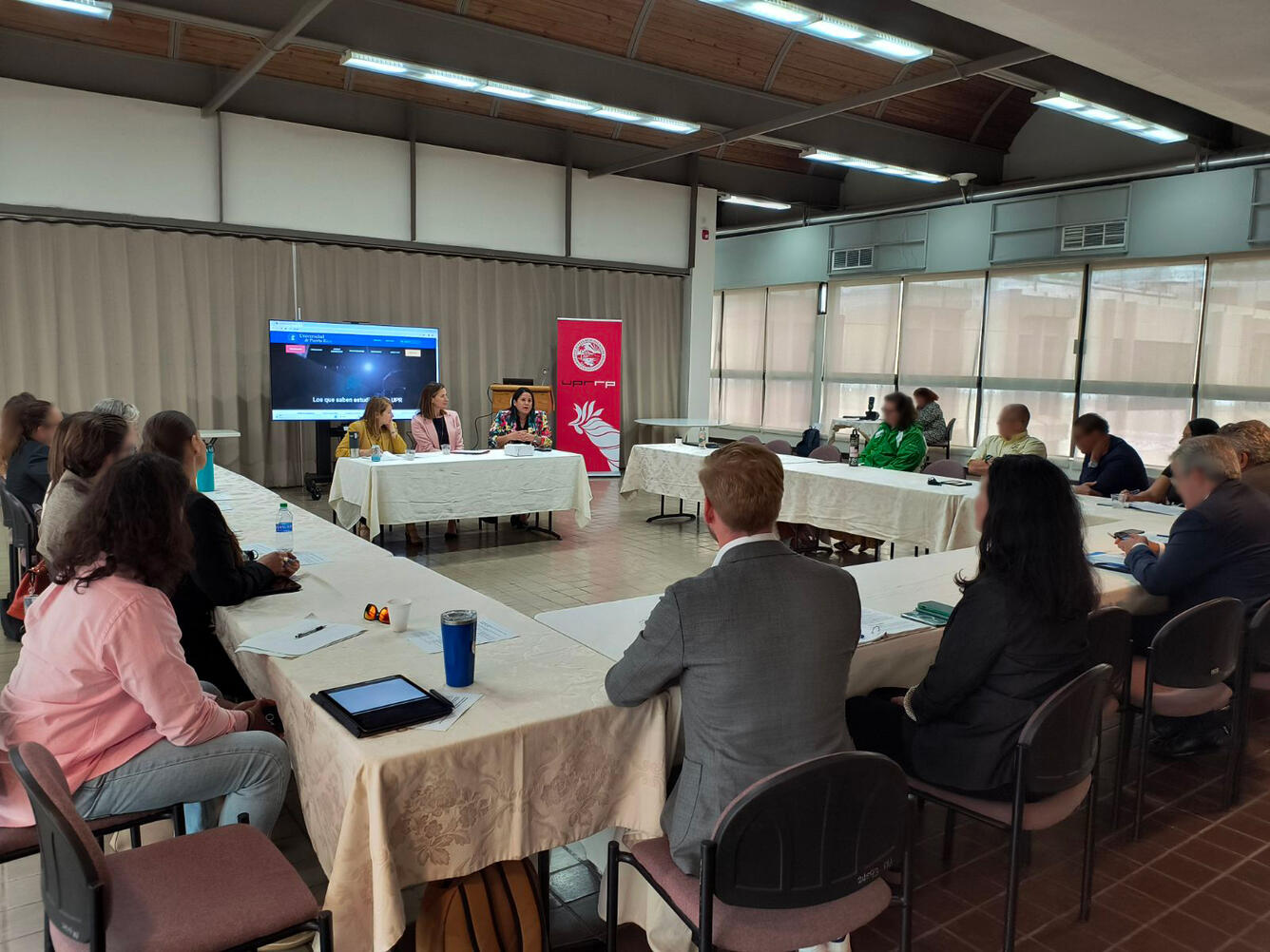 Three women sit at a table with a screen and banner behind them, surrounded by people sitting around a square of tables.