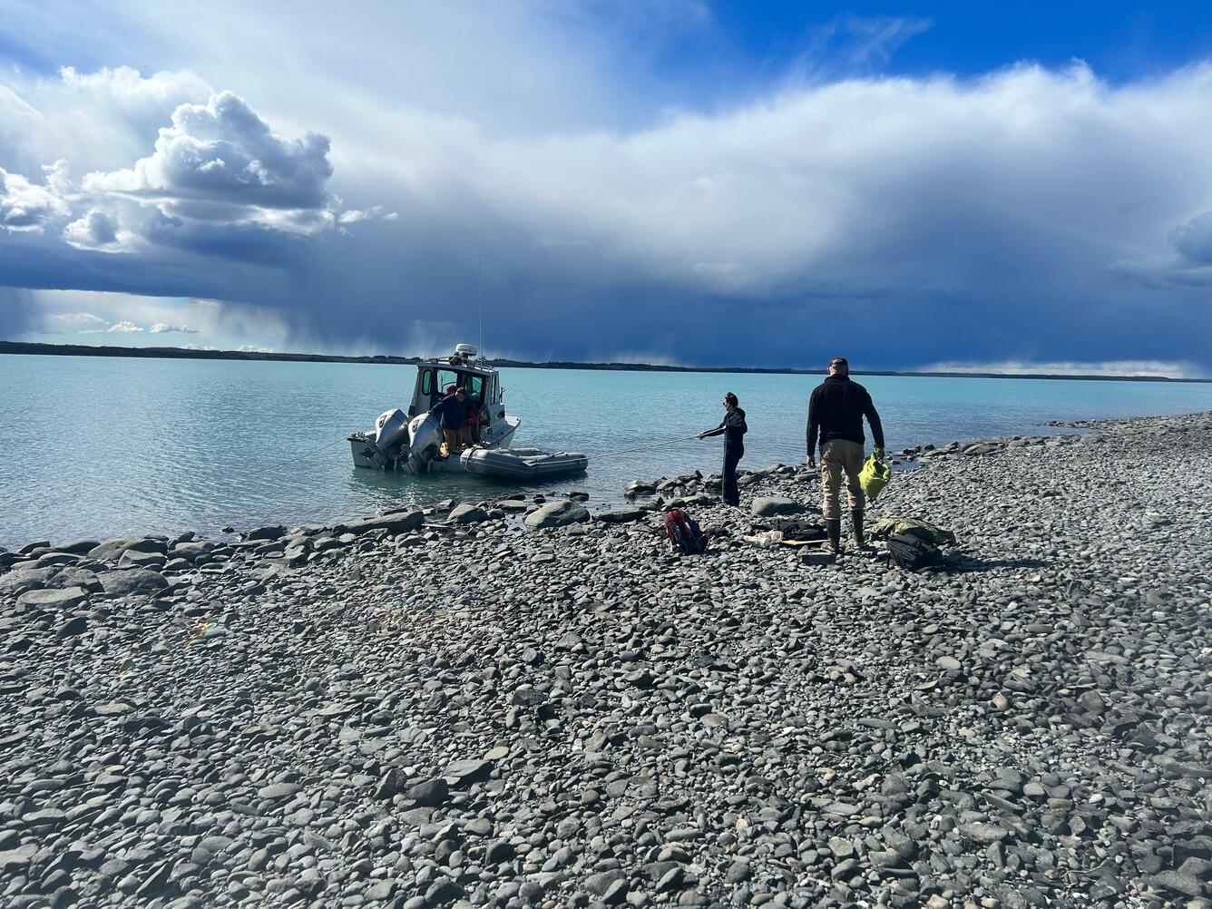 two people stand on rocky beach while boat floats in lake