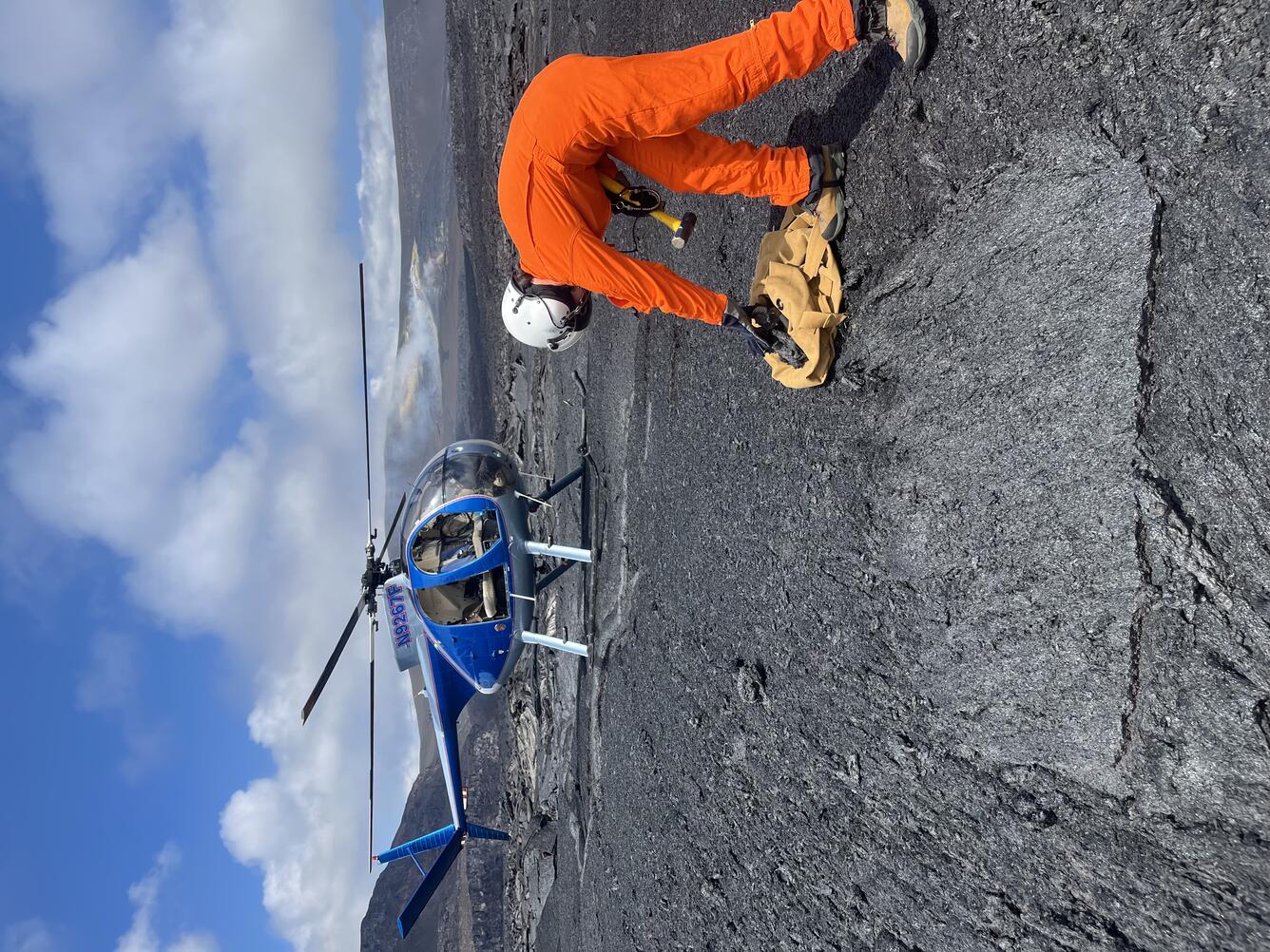 Color photograph of geologist collecting sample of lava flow with helicopter in background