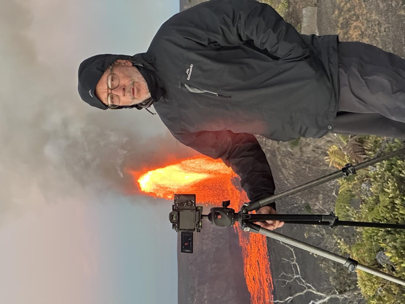 Color photograph of scientist bundled up with camera on tripod next to him and lava fountain erupting in the background