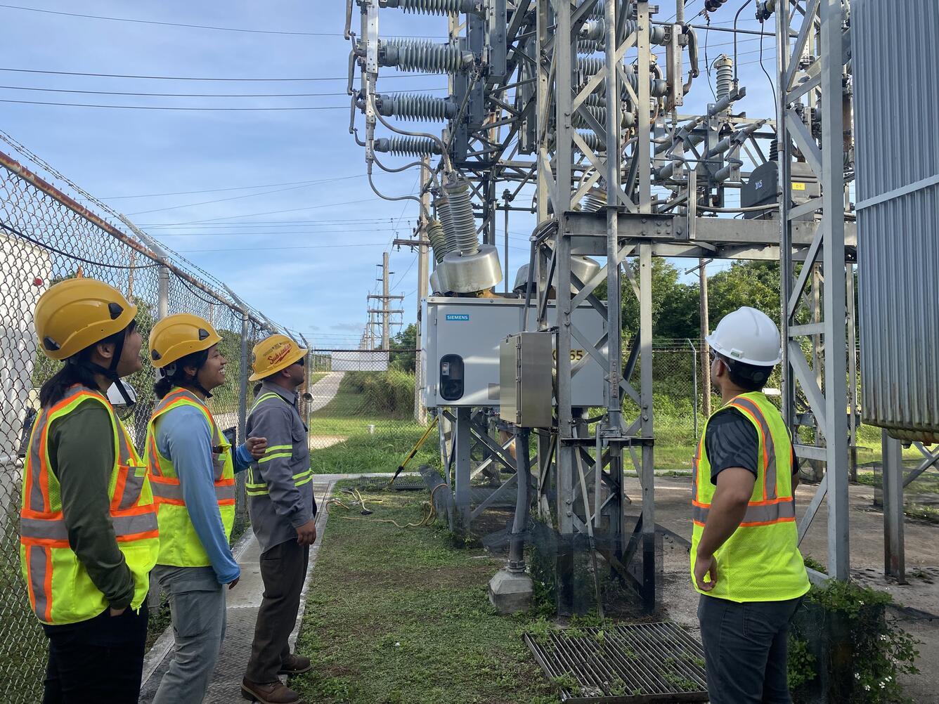 Four workers in hard hats stand within a power substation.
