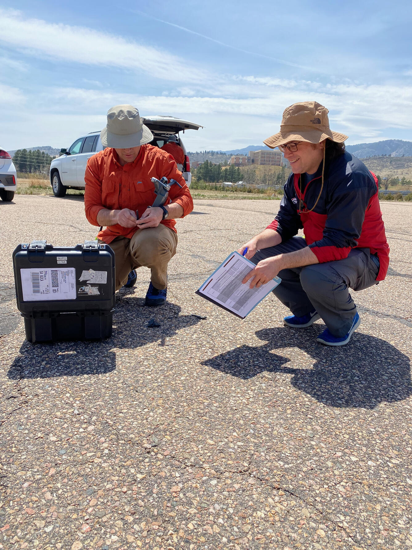 Two people crouched down in parking lot holding some papers and looking in a box