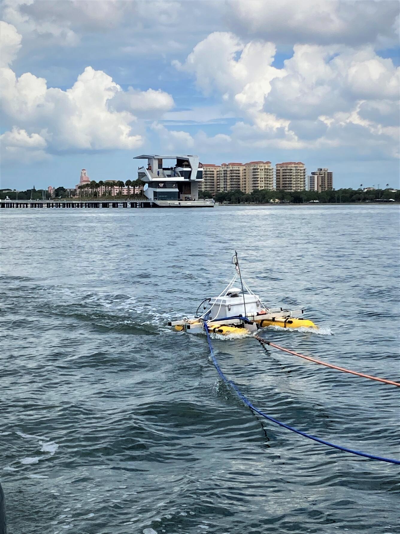 A collection of equipment is mounted on  yellow catamaran in the bay. In the background: a pier & skyline with tall buildings