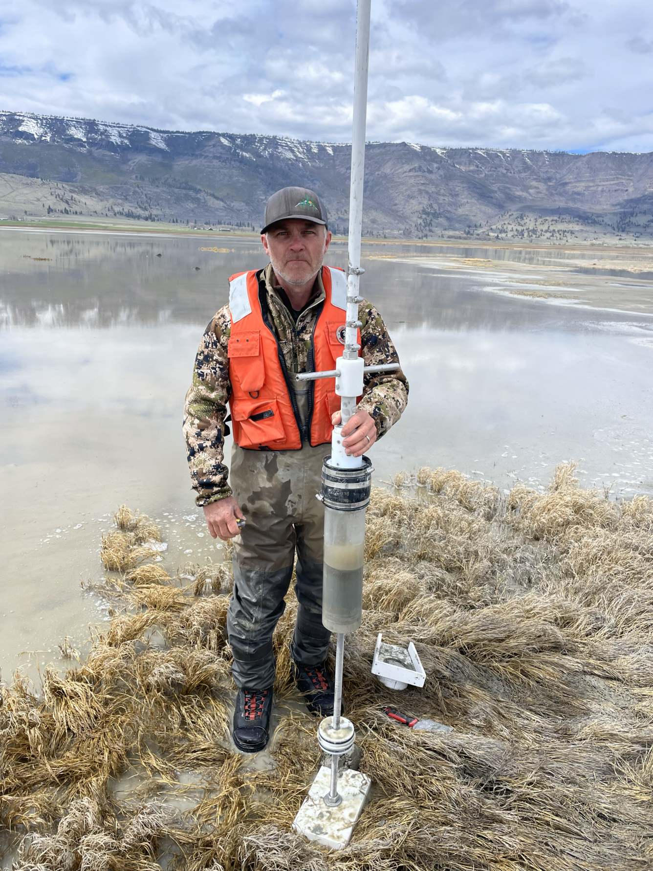 man on shallow lake shore holding a long, clear tube with a handle or frame at the top and a valve or stopper at the bottom