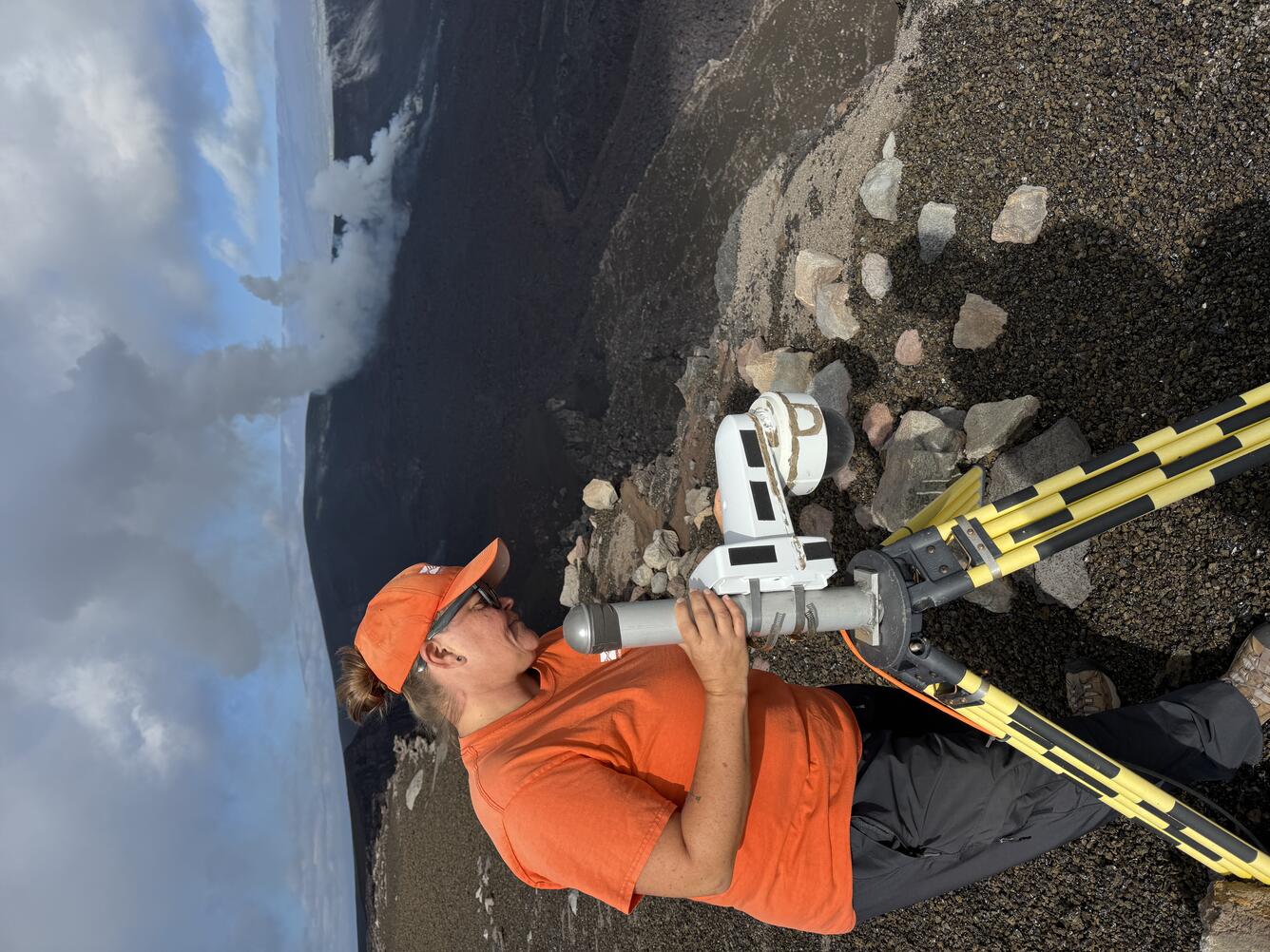Color photograph of field engineer servicing a livestream camera with volcanic vents degassing in the background