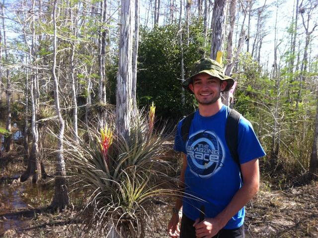 A smiling man in a wide brimmed-hat and a blue shirt stands in a flooded forest.