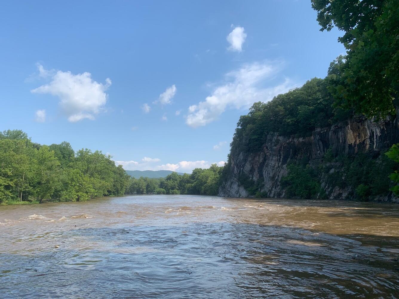 Flooding in the Shenandoah River South Fork. Water is rough and turbid as it flows past a cliffside outcrop.
