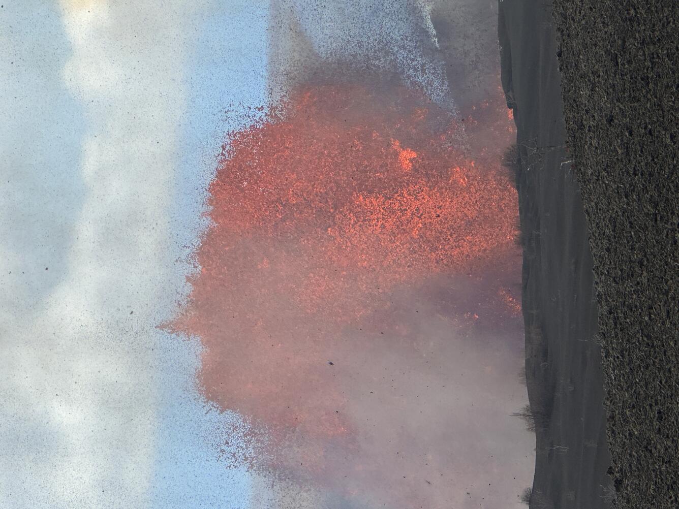 Color photograph of lava fountain during the day time with crater rim in the foreground