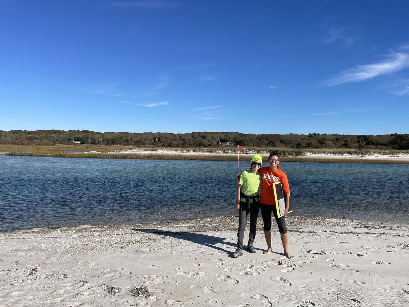 Two people standing next to each other on beach, water, marsh, and sky in background