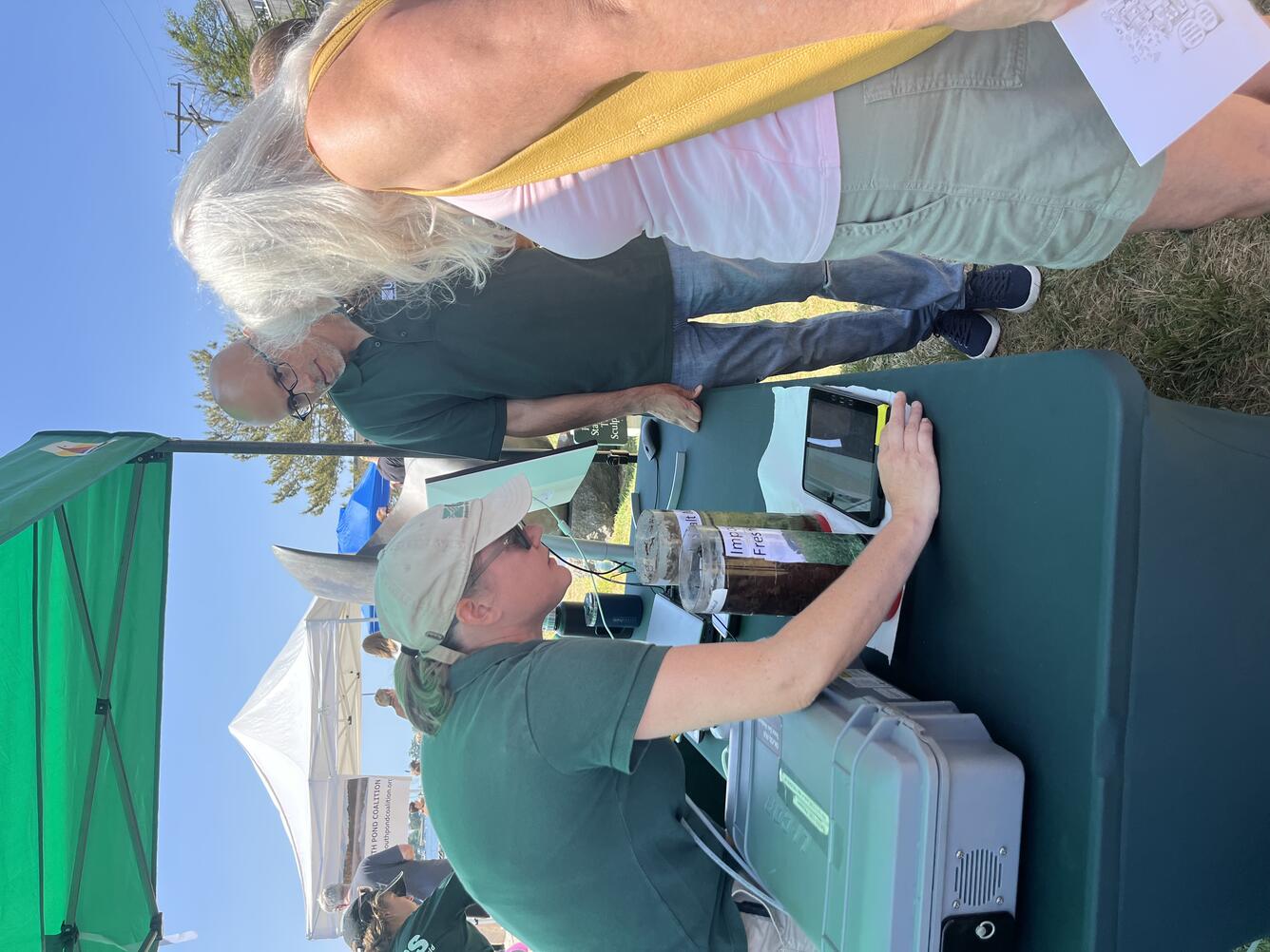 USGS scientist showing visitor something on a tablet on a table outside under a tent