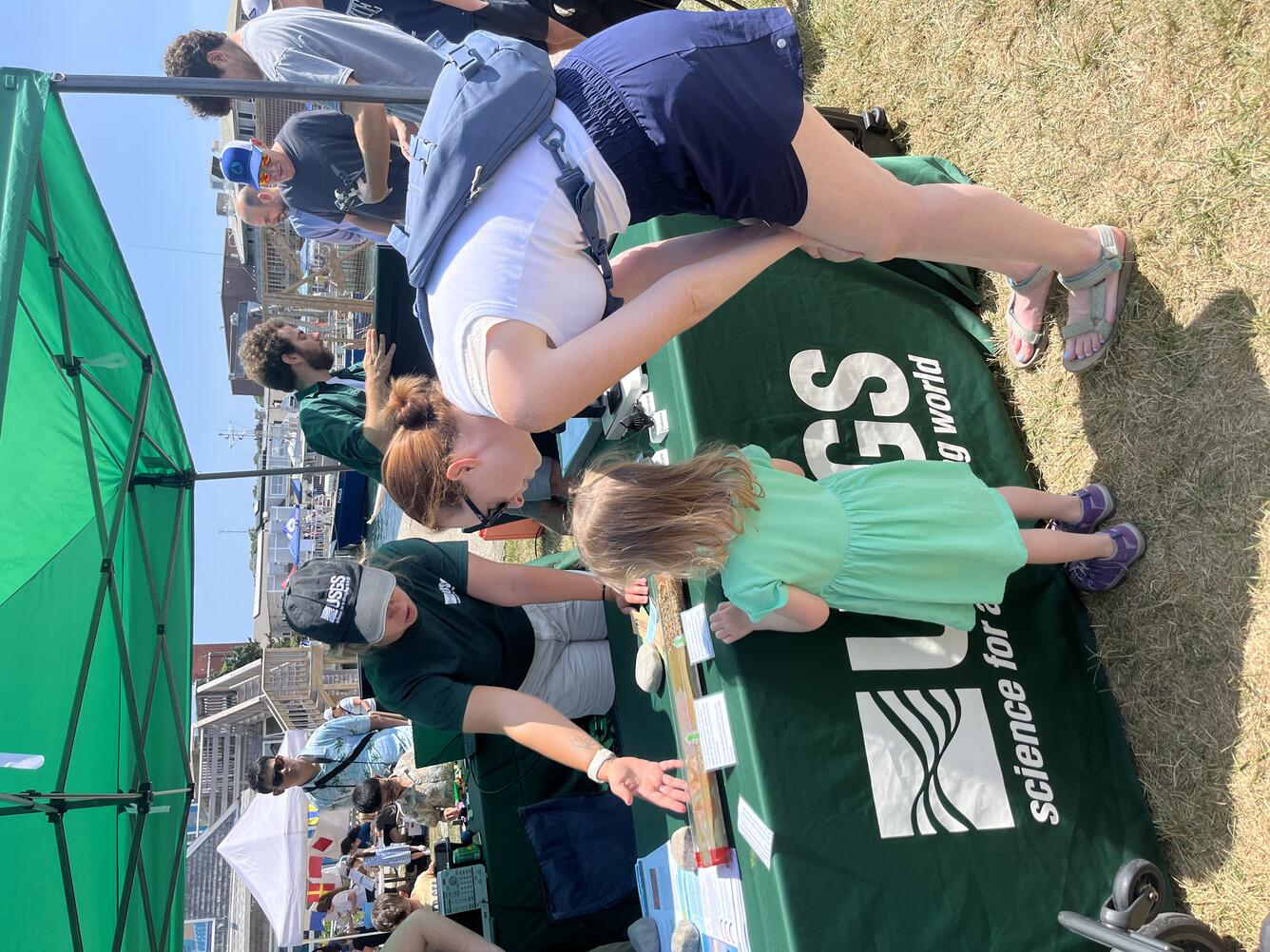 woman showing adult and child a replica sediment core on table outside under green tent