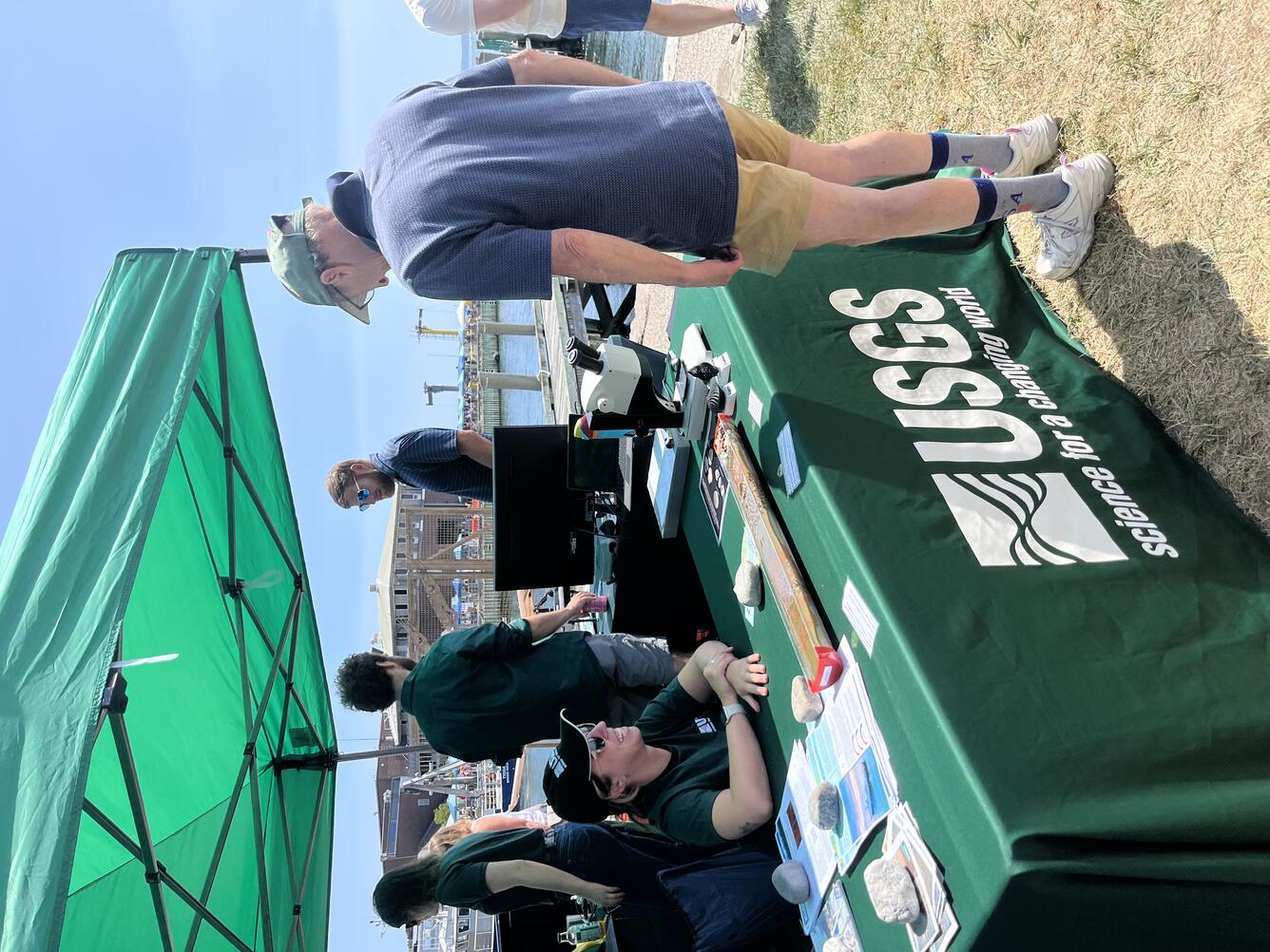 Person sitting at USGS booth under tent outside, smiling at person visiting the booth