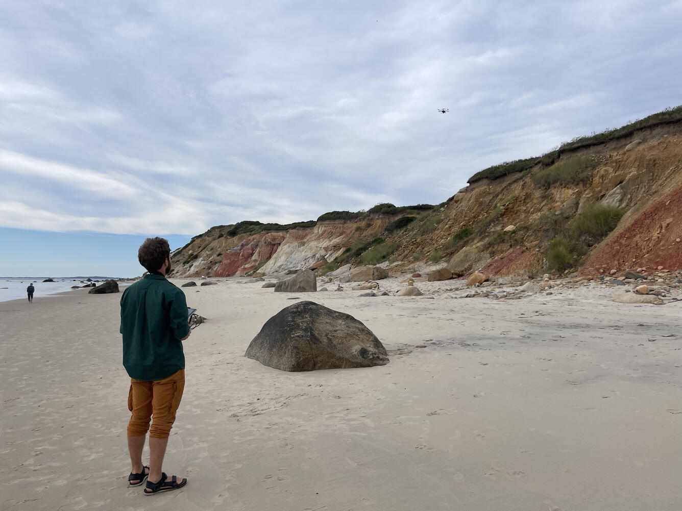 person standing on sandy beach with control in hand looking at drone in the sky over cliffs