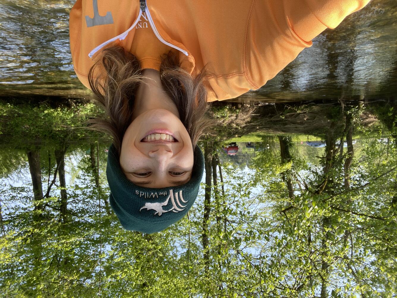 A woman in a green beanie and an orange shirt is smiling and standing in front of a small stream.