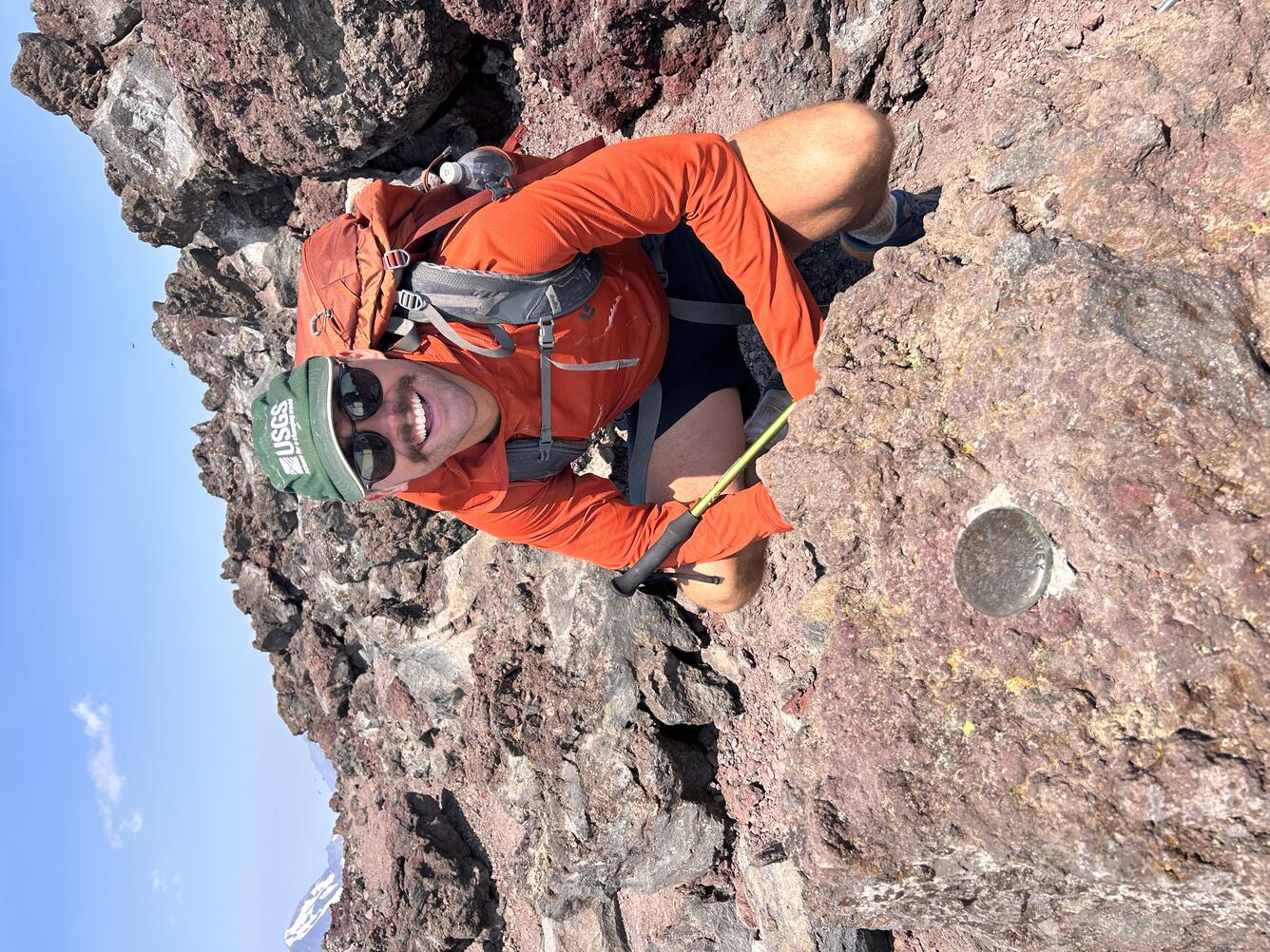 USGS Scientist wearing a USGS logo hat and standing next to a survey monument. 