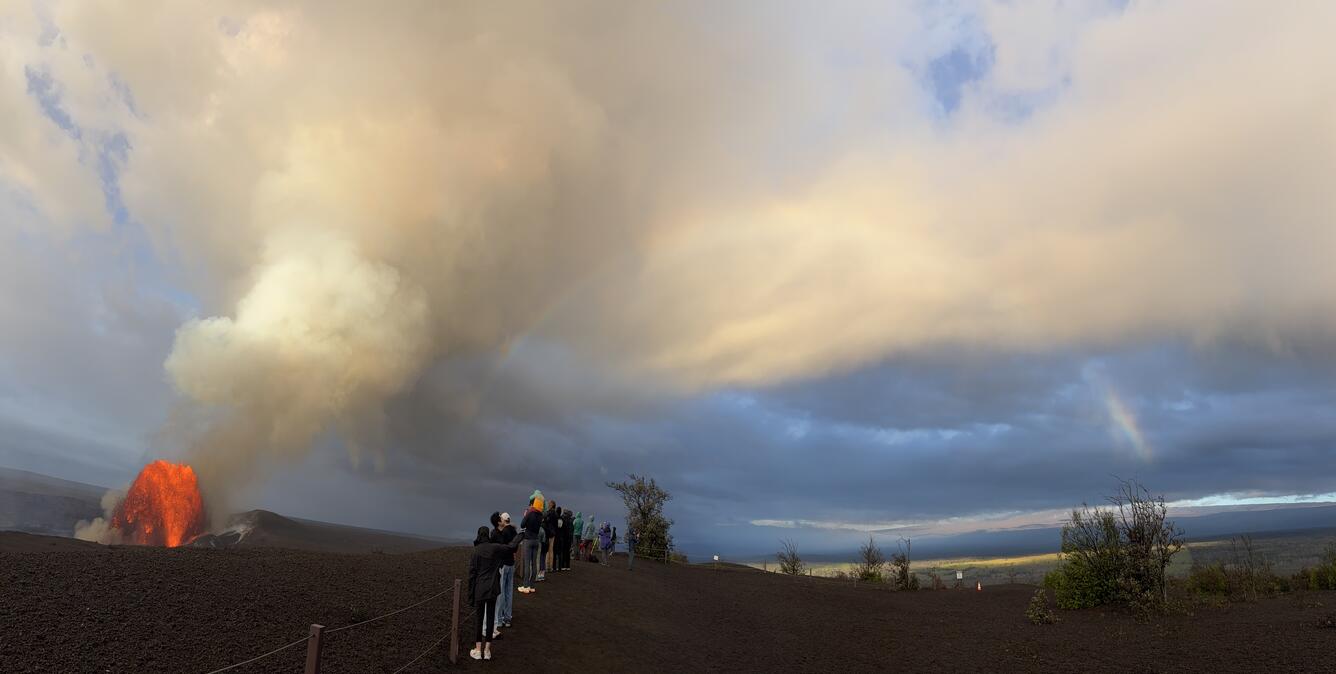 Color photograph of lava fountain in crater, visitors and rainbow on the rim