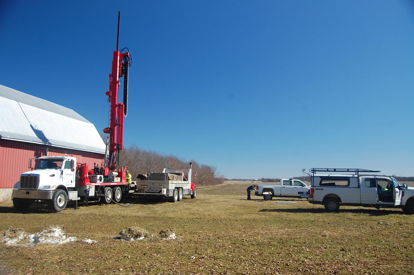 Several vehicles and a large underground drilling rig with a few people in a field beside a building. 