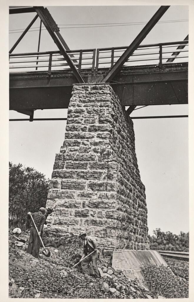 Black and white photo of men digging at the foot of an old bridge