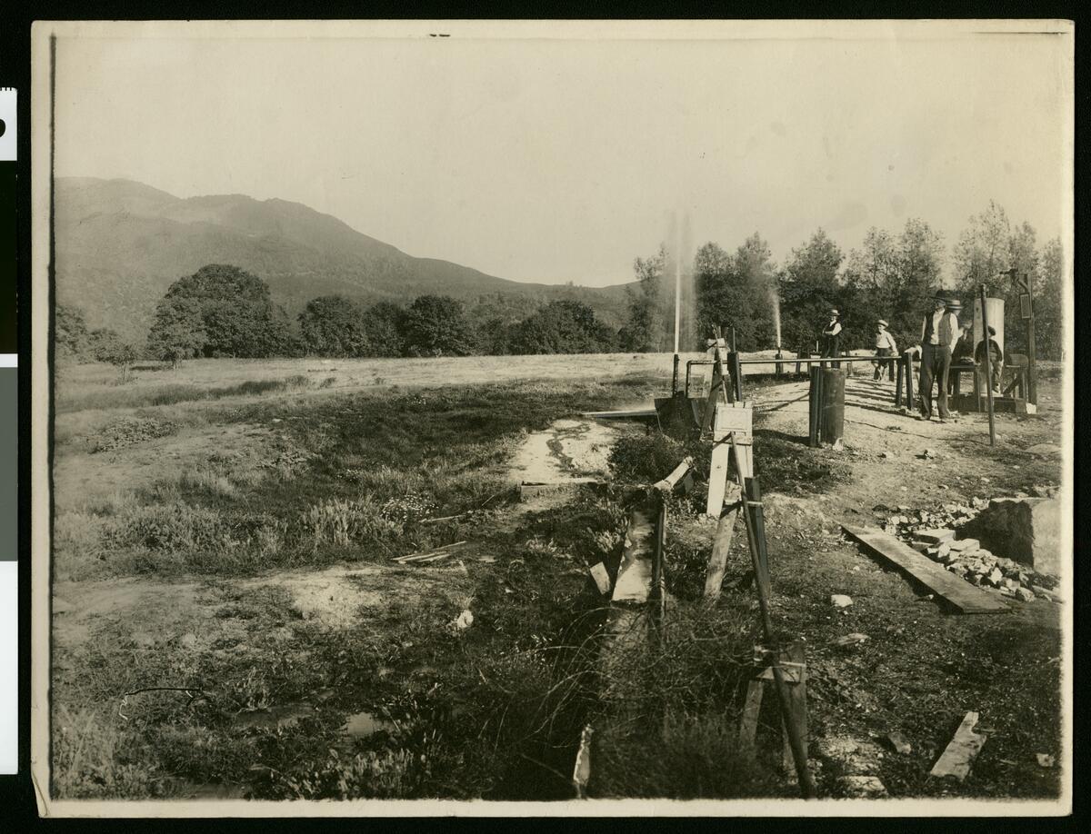 A sepia-toned historical photograph shows an open field with people standing near a row of wooden-framed well structures and pipes. A thin jet of gas or water sprays upward from two of the wells. Forested hills and a mountain rise in the background. 