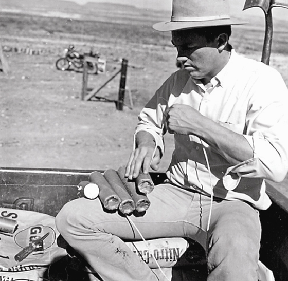 Man in white button-down shirt sitting on bags of explosives, assembling dynamite sticks used as detonator