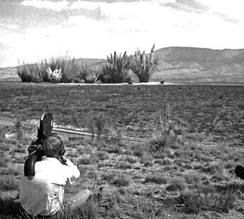 A cameraman (foreground) films small explosions in the distance, which are part of the creation of a crater field
