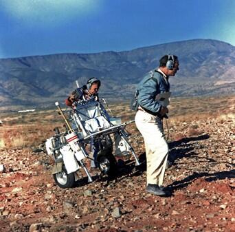 Two men wearing jackets and headsets pulling a piece of equpment up a moderate slope in the desert southwest