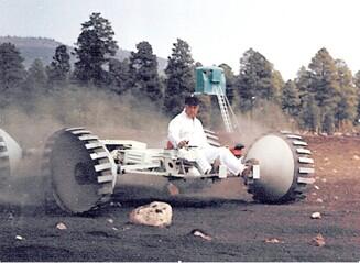 Early lunar roving vehicle prototype with large wheels and an open seat, being driven by a man in a white jumpsuit