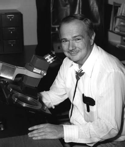Gene Shoemaker wearing a white button-up shirt and a bolo tie, sitting at a microscope and looking at the camera