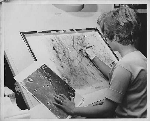 A woman with short hair sits at an inclined desk, adding detail to a map of the lunar surface with an airbrush