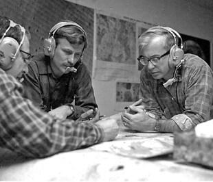 Three men with headsets are hovering over maps and appearing to discuss observations