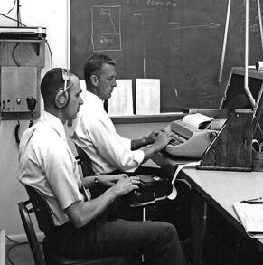 Two men in white shirts sitting at typing machines: a typewriter and a stenograph