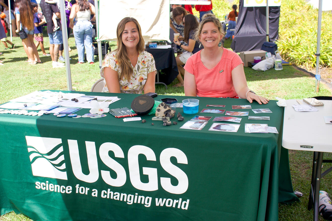 Two women sit at a USGS booth table at an outdoor event. 