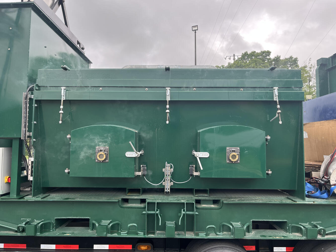 Rear view of a green industrial trailer or tank body, mounted on a flatbed truck chassis. The tank has two arched access doors with round latch handles and gauge/valve fittings, flanked by turnbuckle tensioners along the top edge. Cables run between the two door latches. Below, the flatbed frame shows tie-down hooks and chain slots. 