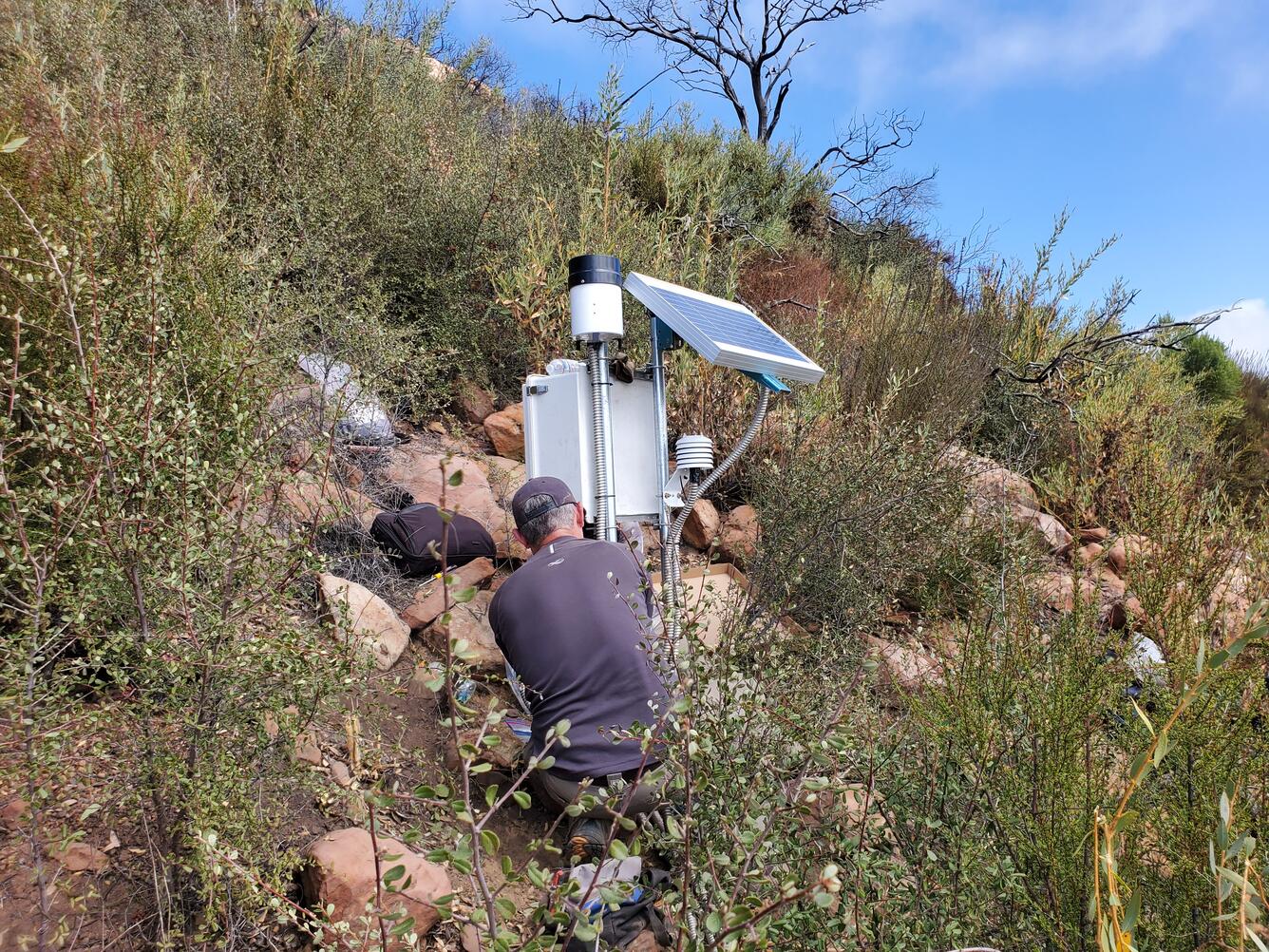 Person kneeling next to equipment surrounded by rocks and shrubs on a hillslope