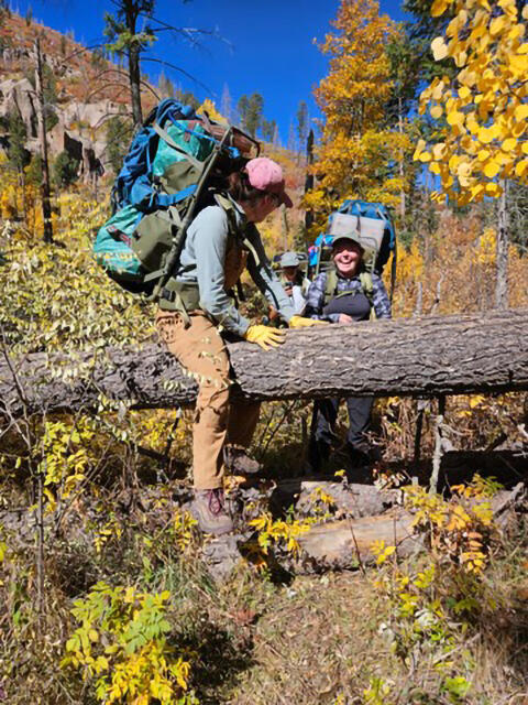 Field station member climbs over log with a full backpack while another field station member approaches