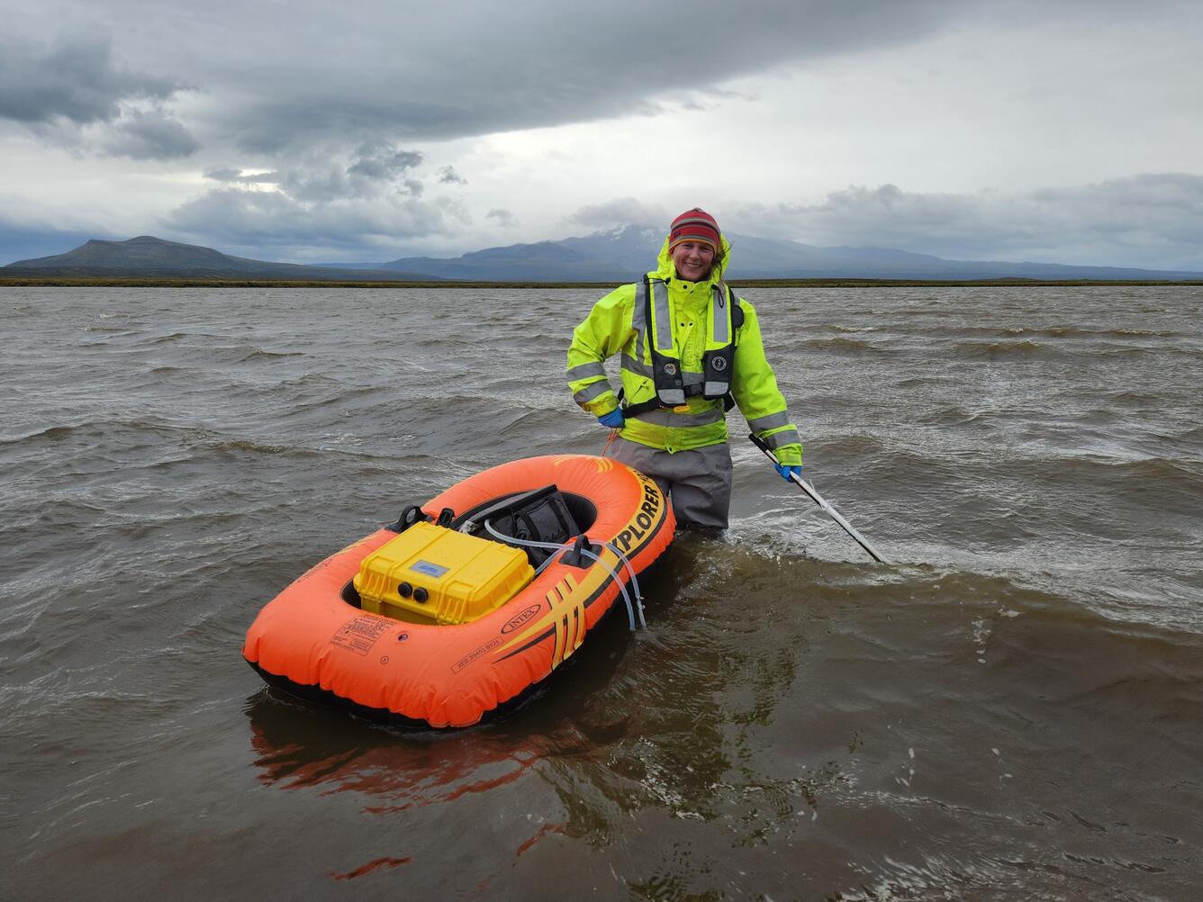 Scientist in bright yellow jacket smiles while standing in thigh-deep water with an orange inflatable boat and filter device