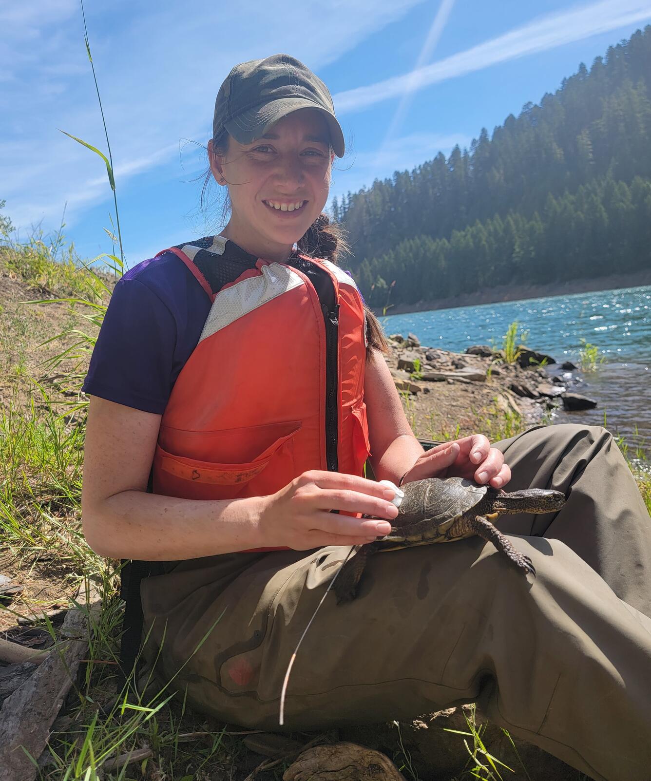 photo of a person wearing an orange life jacket sitting next to a lake holding a turtle