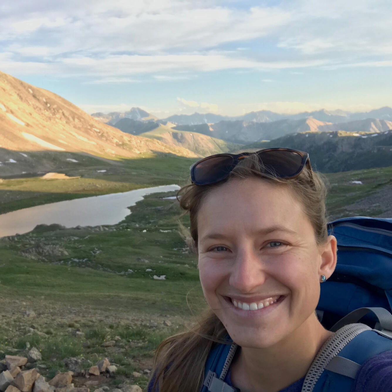 woman with blonde hair wearing a blue backpack standing near a river and mountains on a partly sunny day