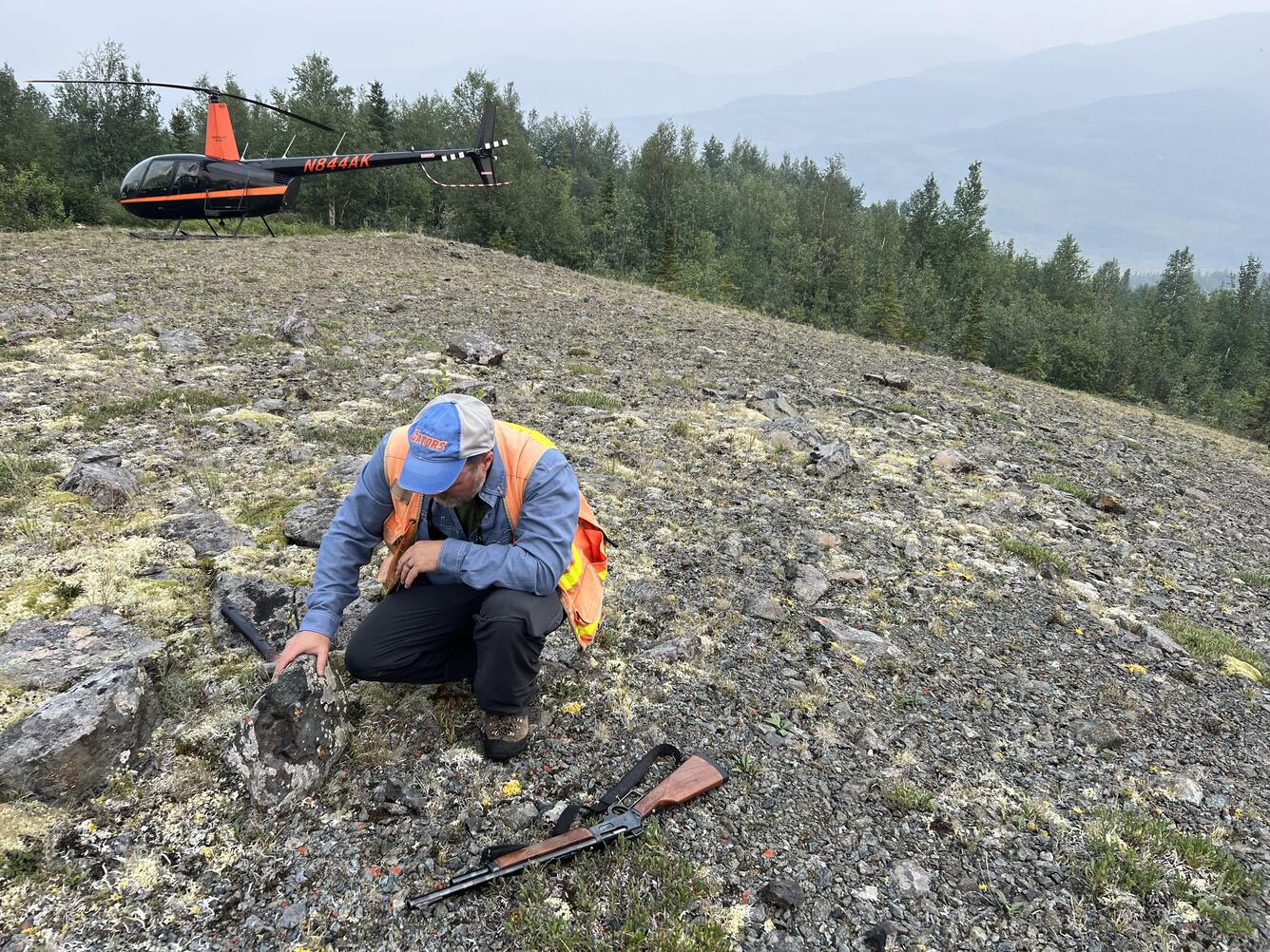 Male geologist, kneeling with hand on rock with rifle on ground, and helicopter and evergreen trees in background.