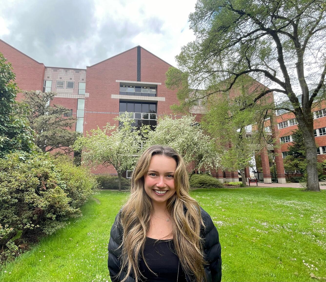 A young woman with blonde hair stands in a park while smiling for her picture.