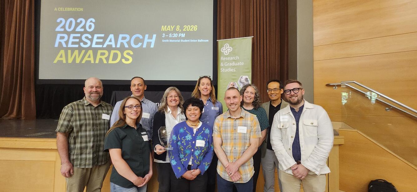10 people stand and smile in front of a light wood stage with a projector screen saying: 2026 Research Awards