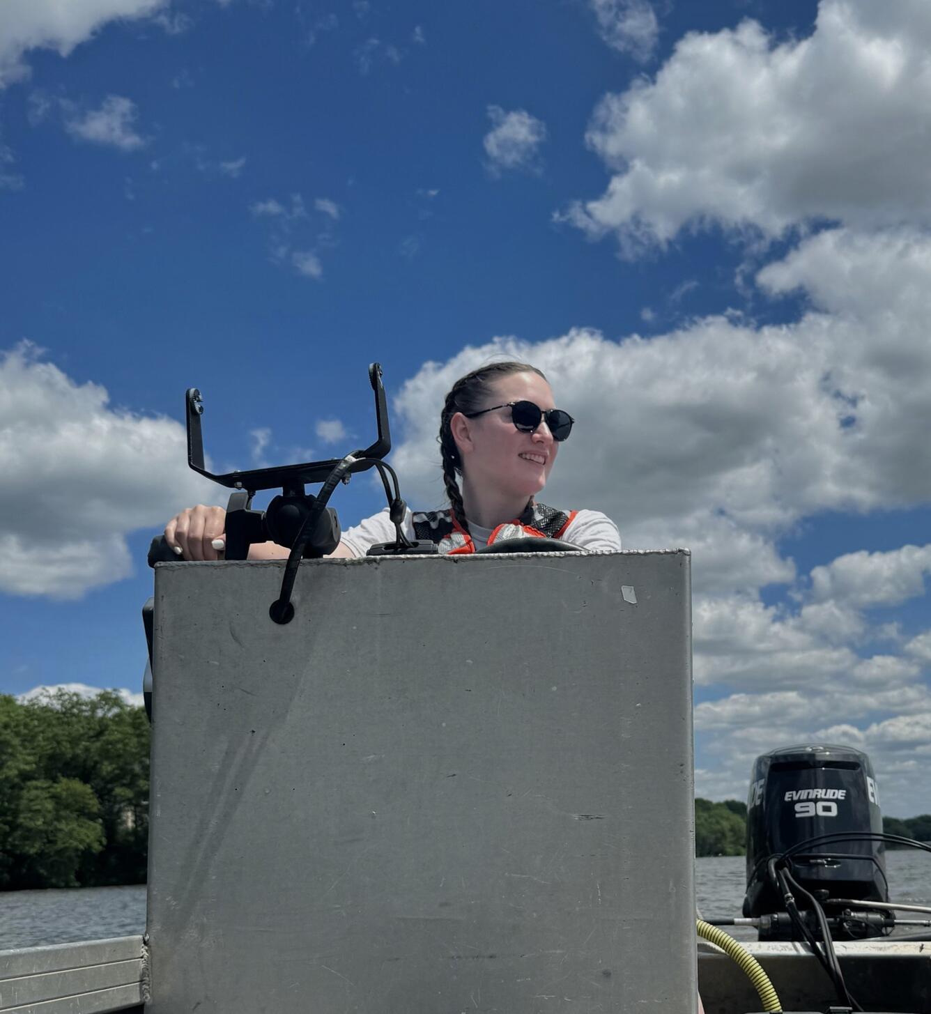 USGS Geographer wearing an orange personal floatation device standing behind the console of a boat against a blue sky.