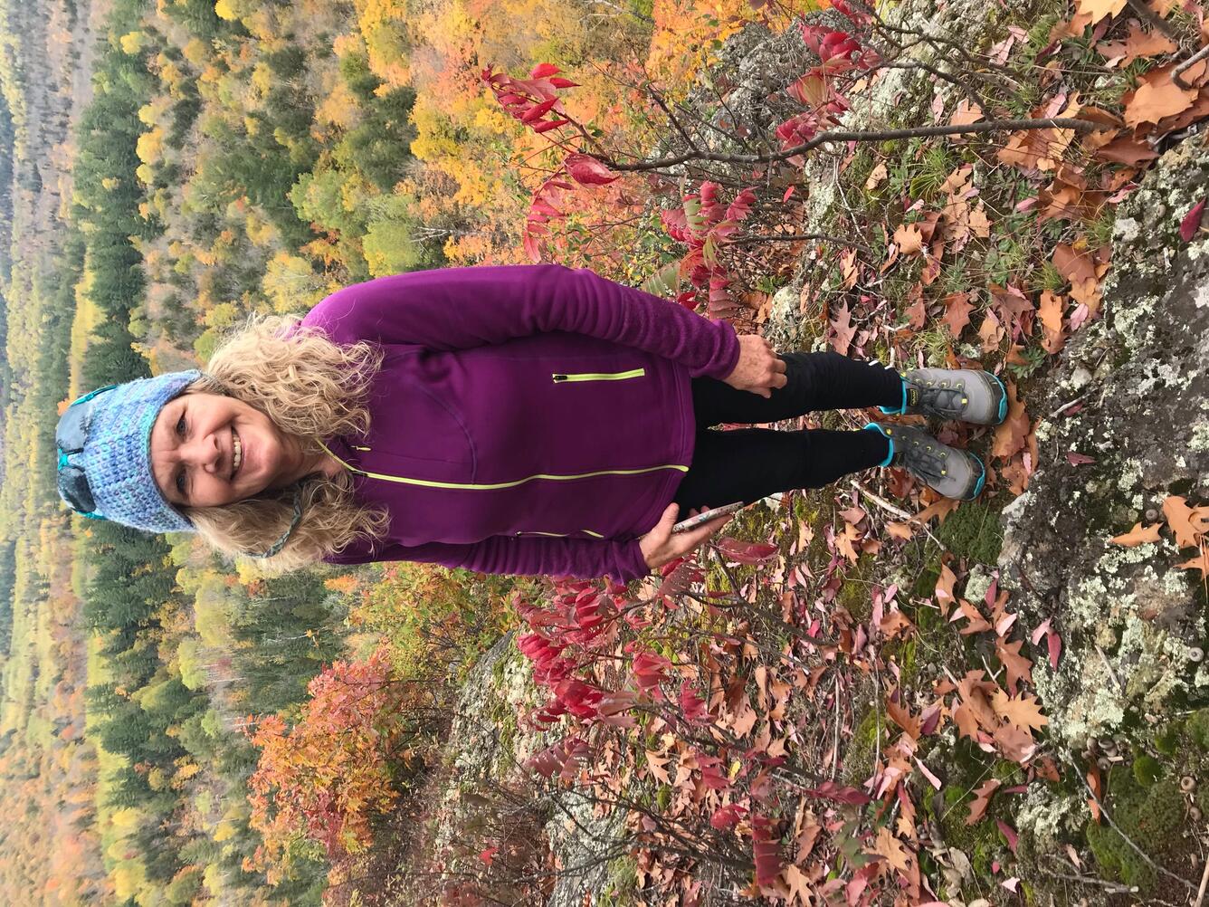Woman in blue hat and burgundy jacket standing on hill covered in autumn colored leaves looking over a valley
