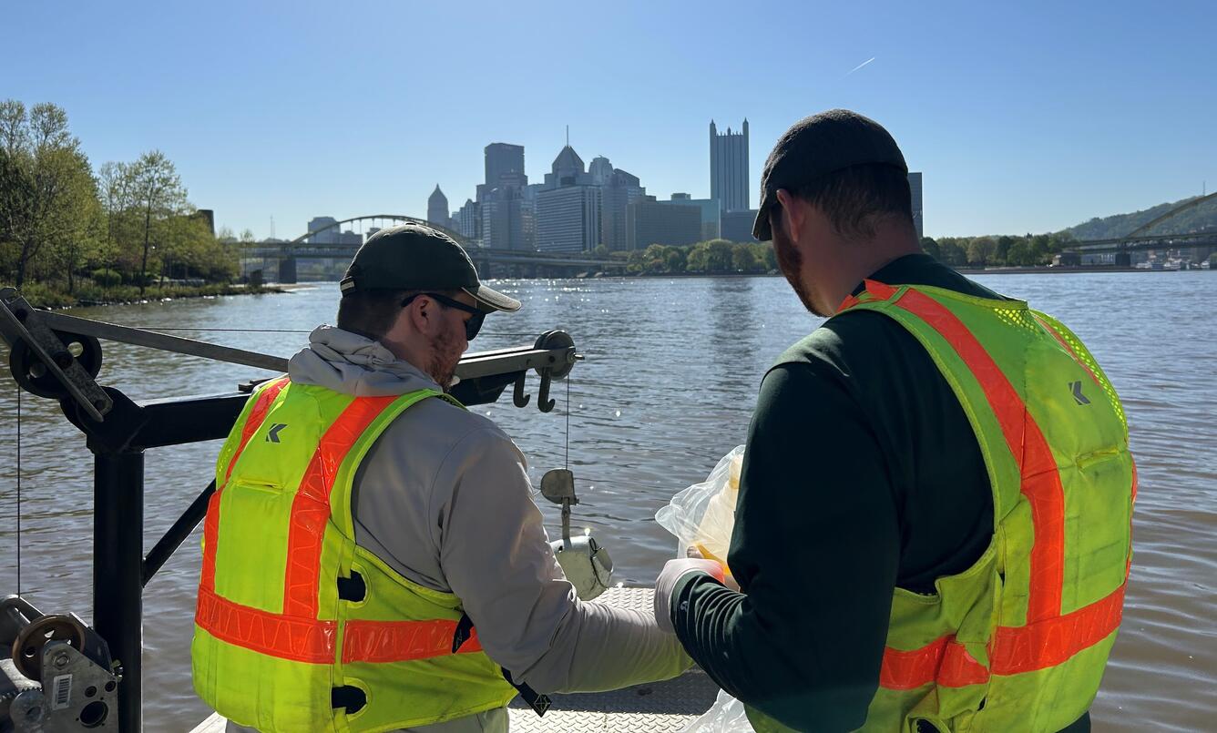 Two people on a boat collecting water samples with downtown Pittsburgh skyline in the background