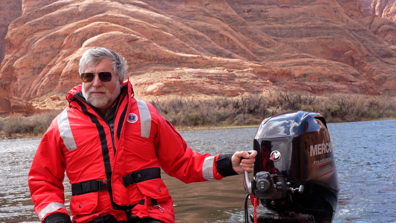 A man with white hair and a beard wears an orange waterproof jacket as he steers a motorboat through red canyons.